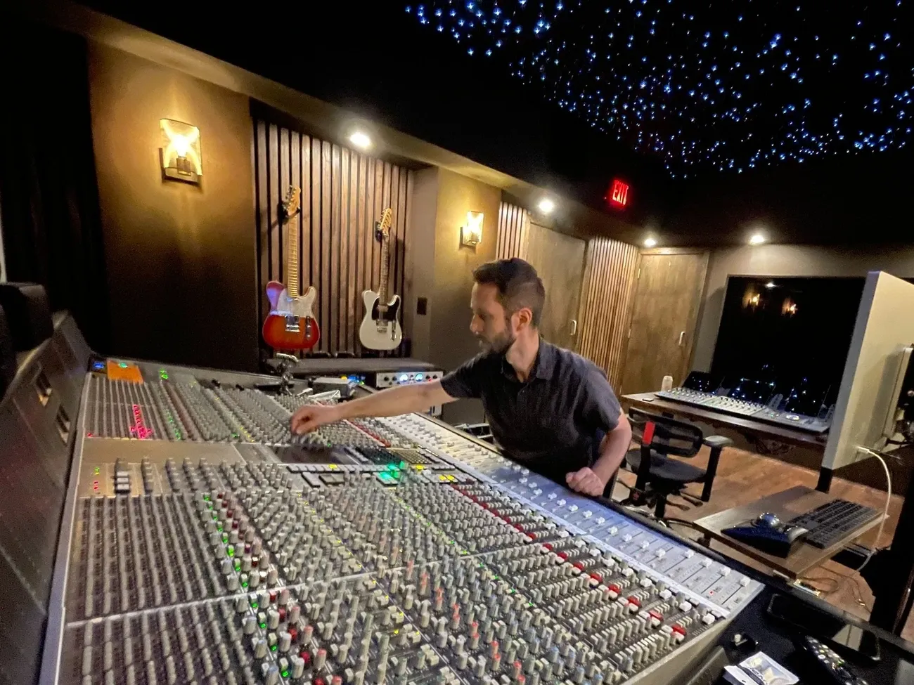 Audio engineer adjusting controls on a large mixing console in a dimly lit recording studio with guitars on the wall.