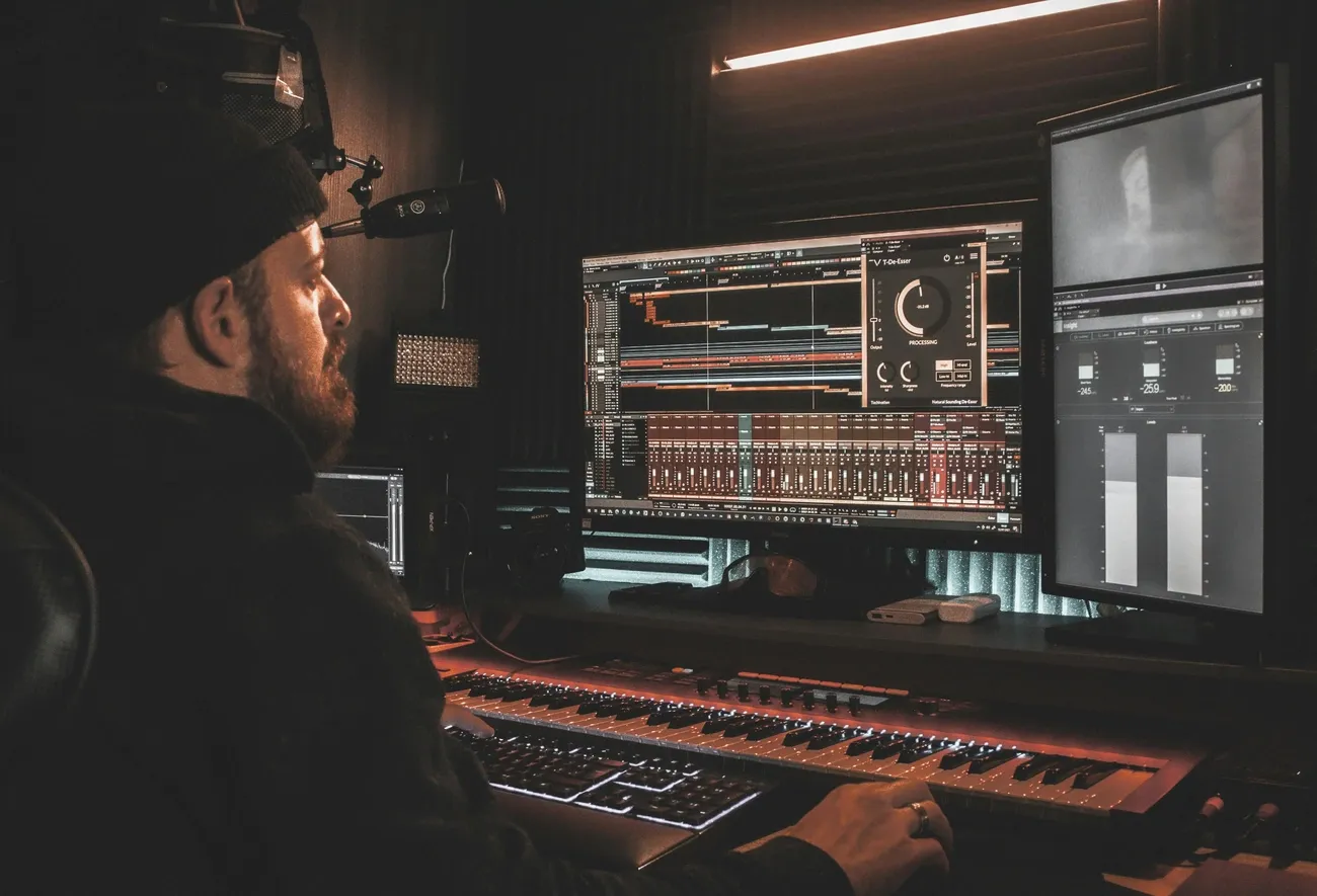 Person working on music production with dual monitors and a MIDI keyboard in a dimly lit studio.