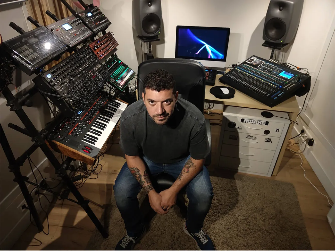 Tech music producer's compact studio with synthesizers, drum machines, mixer, speakers, and computer setup.