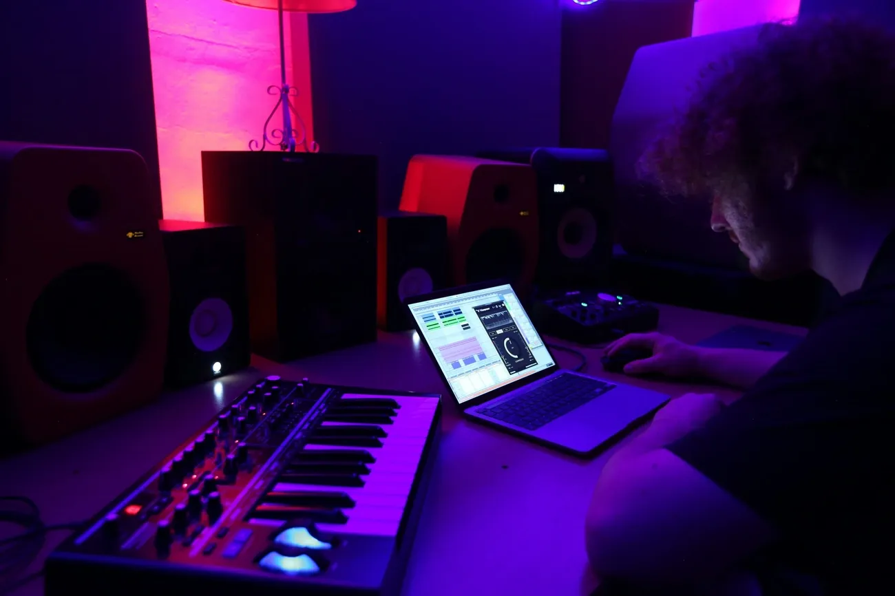 Music producer working on a laptop with a MIDI keyboard and studio monitors in a dimly lit room.