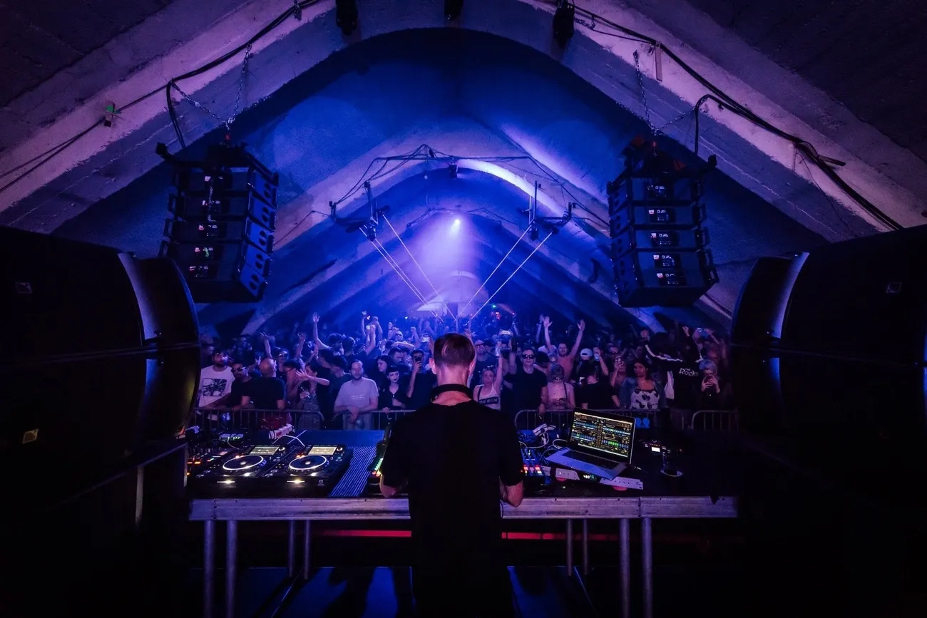 DJ performing techno set on stage with crowd dancing under blue-lit vaulted ceiling in club venue.