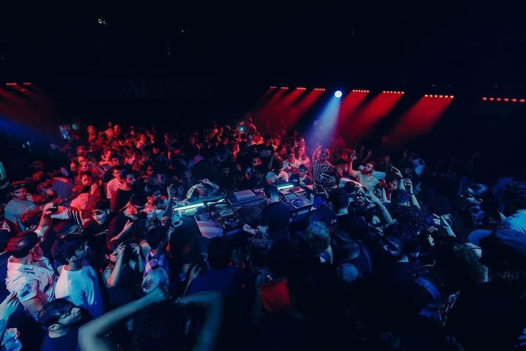 Crowded nightclub scene with a DJ performing under red and blue stage lights during a techno event.
