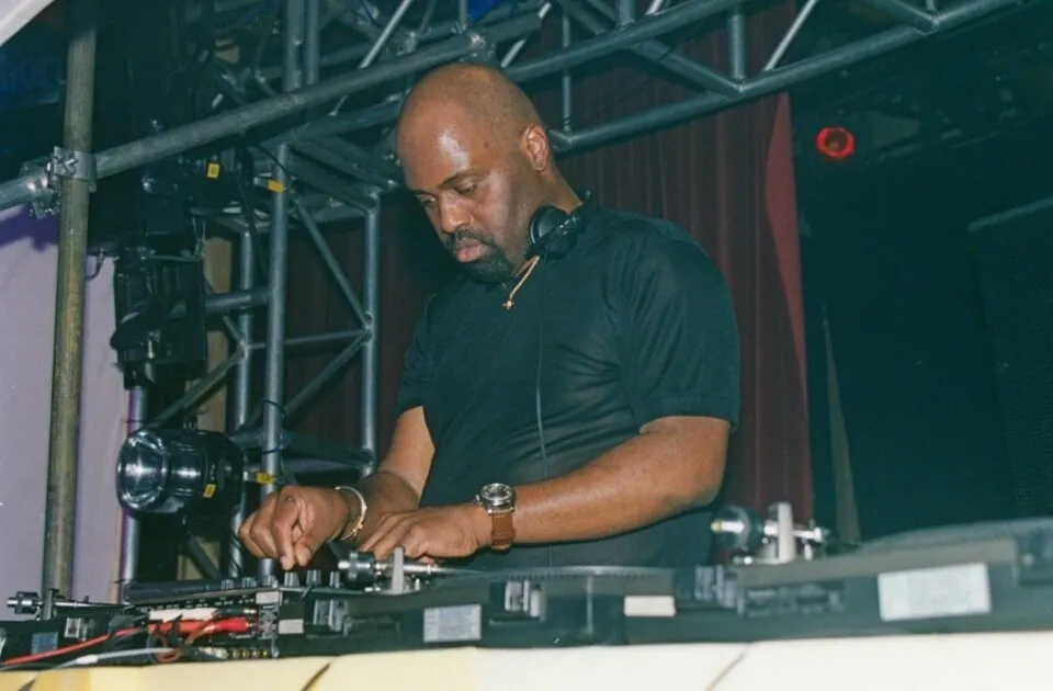 A DJ wearing headphones around his neck performs on turntables at an electronic music event with stage lighting equipment.