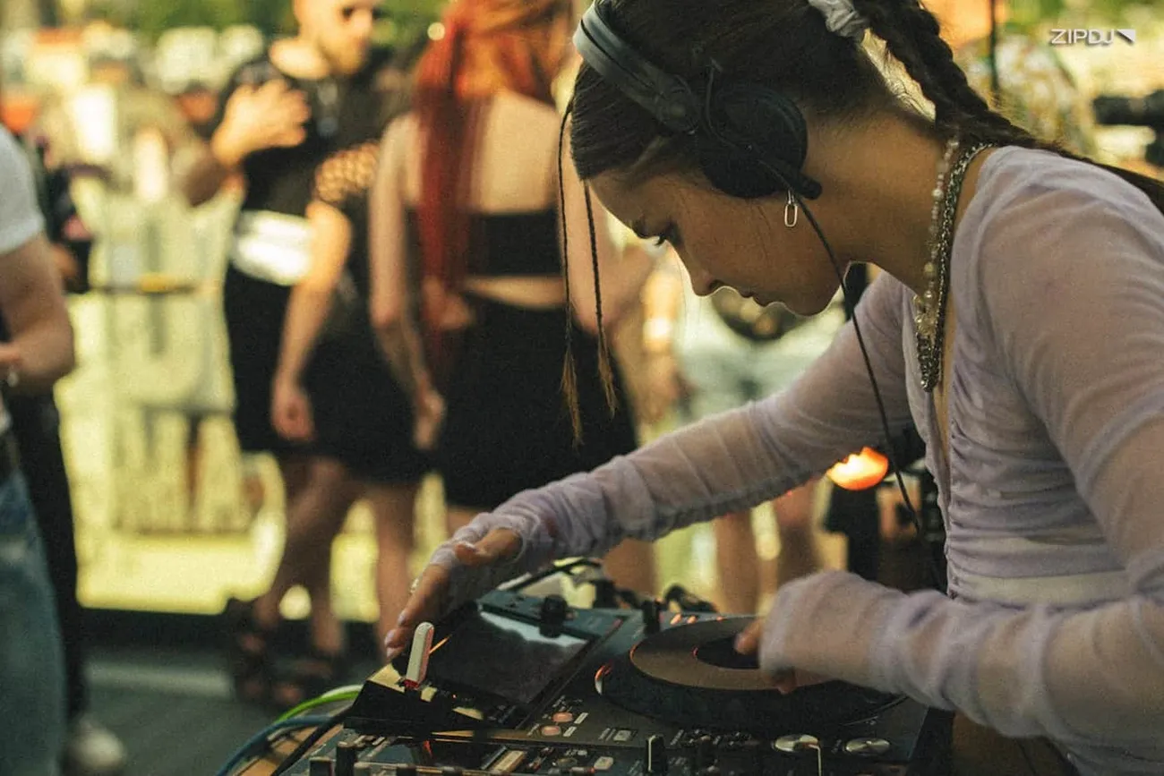 DJ wearing layered necklaces and a long-sleeve top adjusts controls on a DJ mixer at an outdoor event.