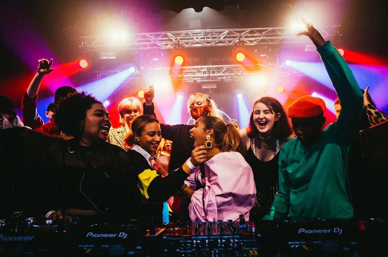 A lively crowd dances and raises hands around DJ equipment under colorful stage lights in a club setting.