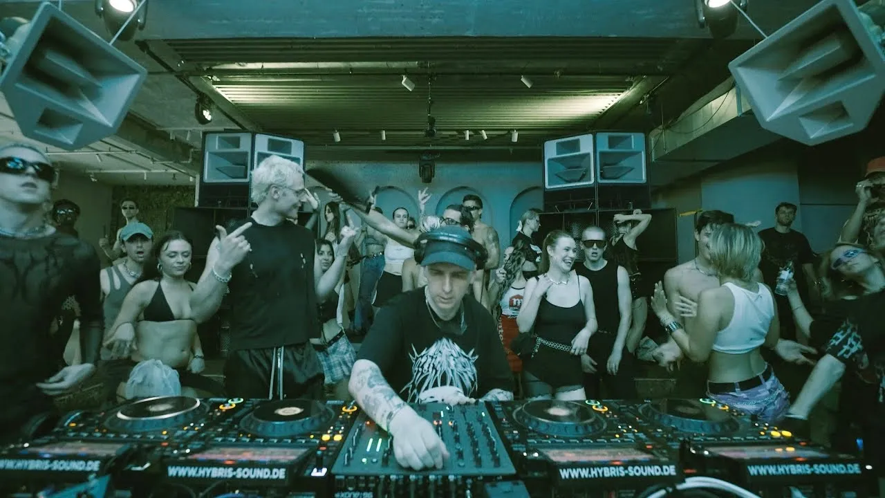 DJ performing at a crowded indoor rave party with people dancing and socializing around the sound equipment.
