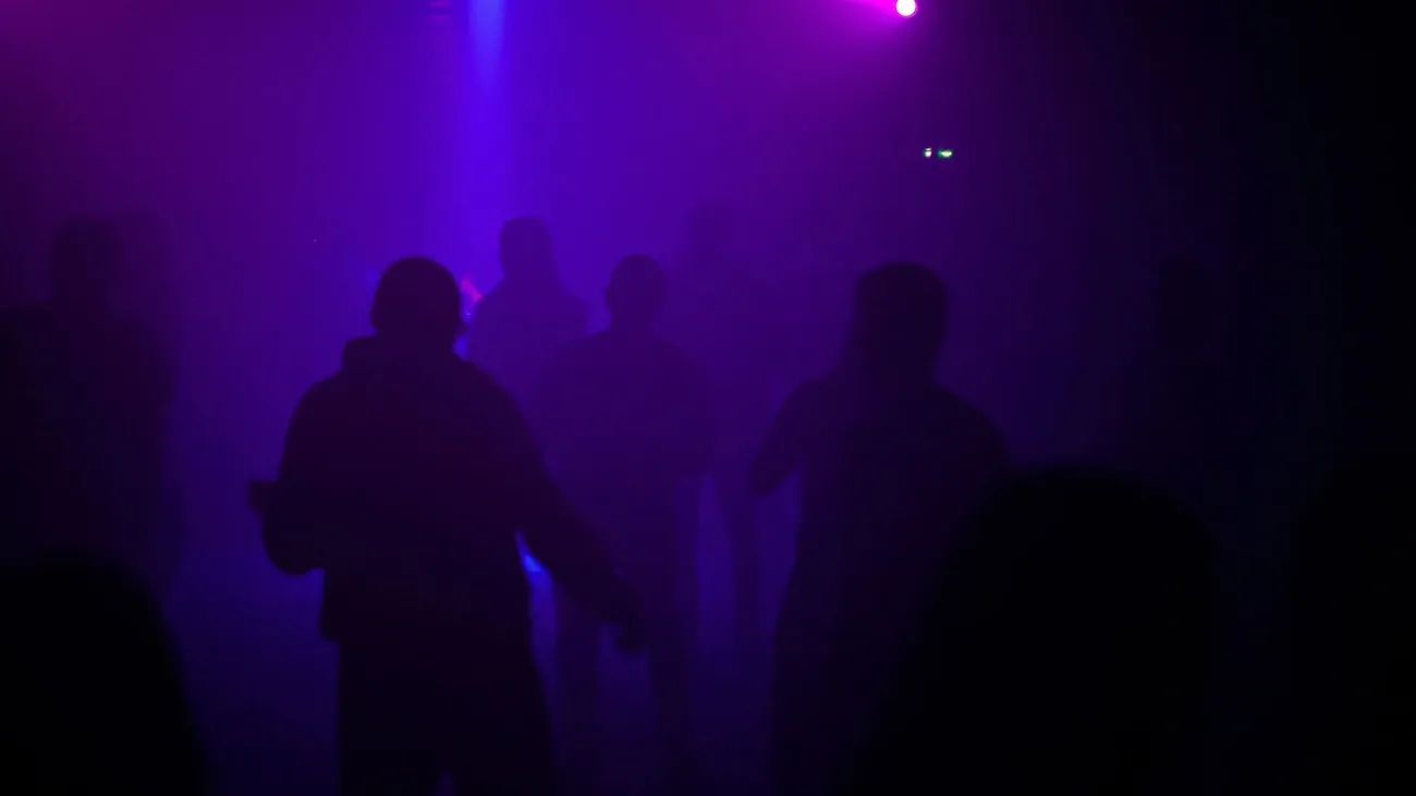 Silhouettes of people dancing in a dark, purple-lit techno club filled with haze and ambient light.
